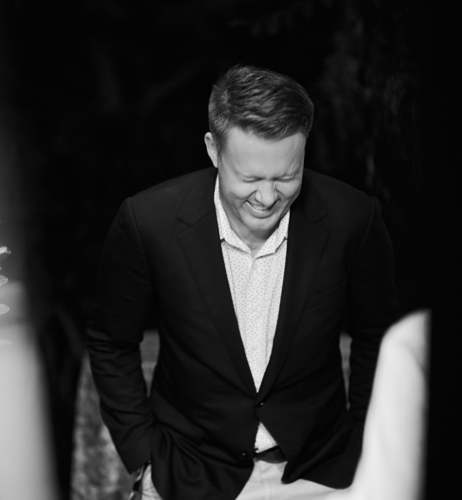 Candid black and white photo of a man in a suit jacket laughing joyfully.