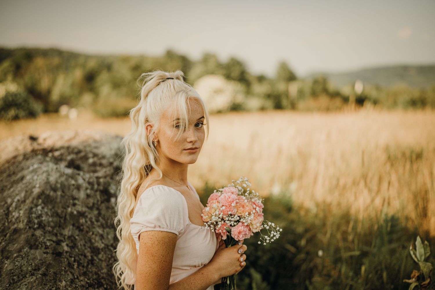 Blonde woman in white dress holding pink flower bouquet posing near large rock in golden field.