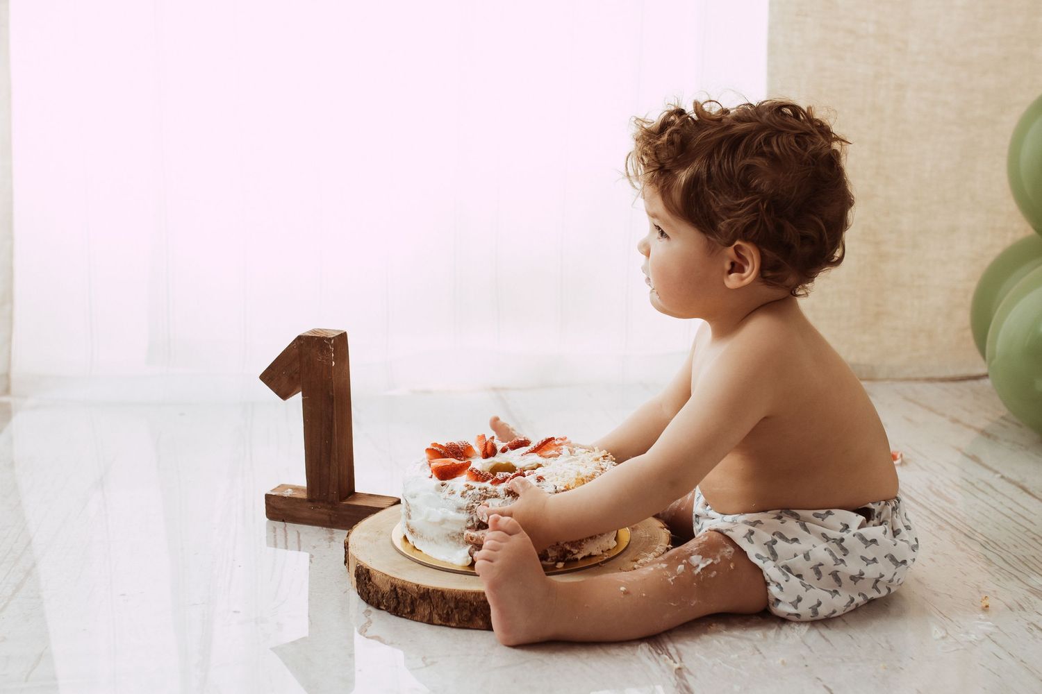 A small child in a cloth diaper sits next to a wooden number one and plate during their first birthday celebration.