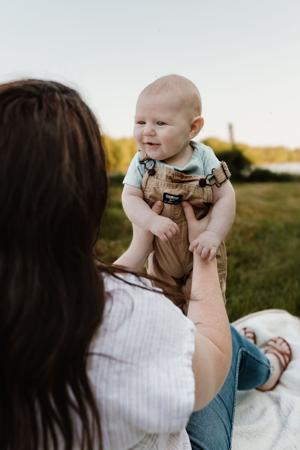 A baby in casual clothes smiles while being lifted in an outdoor setting.