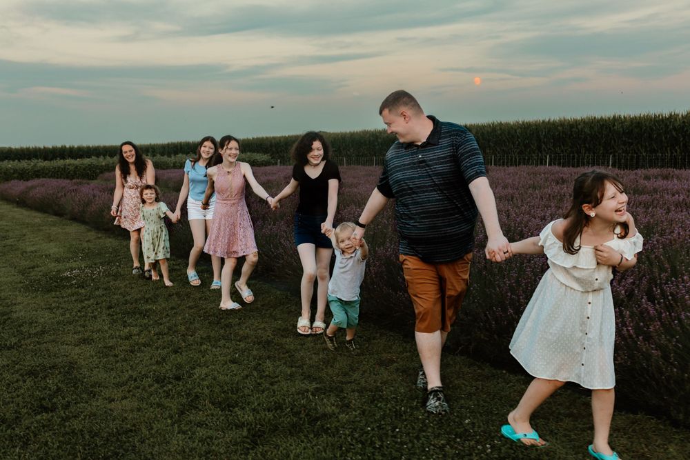 A group walks in single file holding hands along a grassy path at sunset.