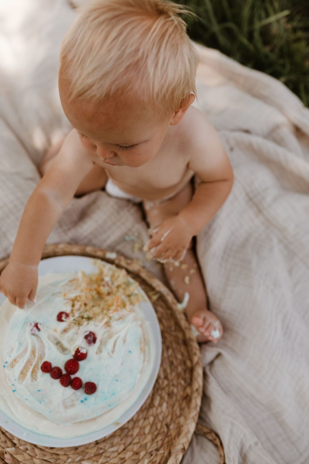 A baby reaching for a decorated birthday cake with raspberries on a woven placemat.