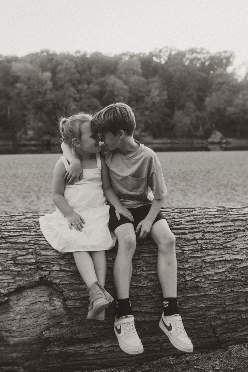 Two children sit together on a fallen log by a lake in a vintage-style black and white photo.