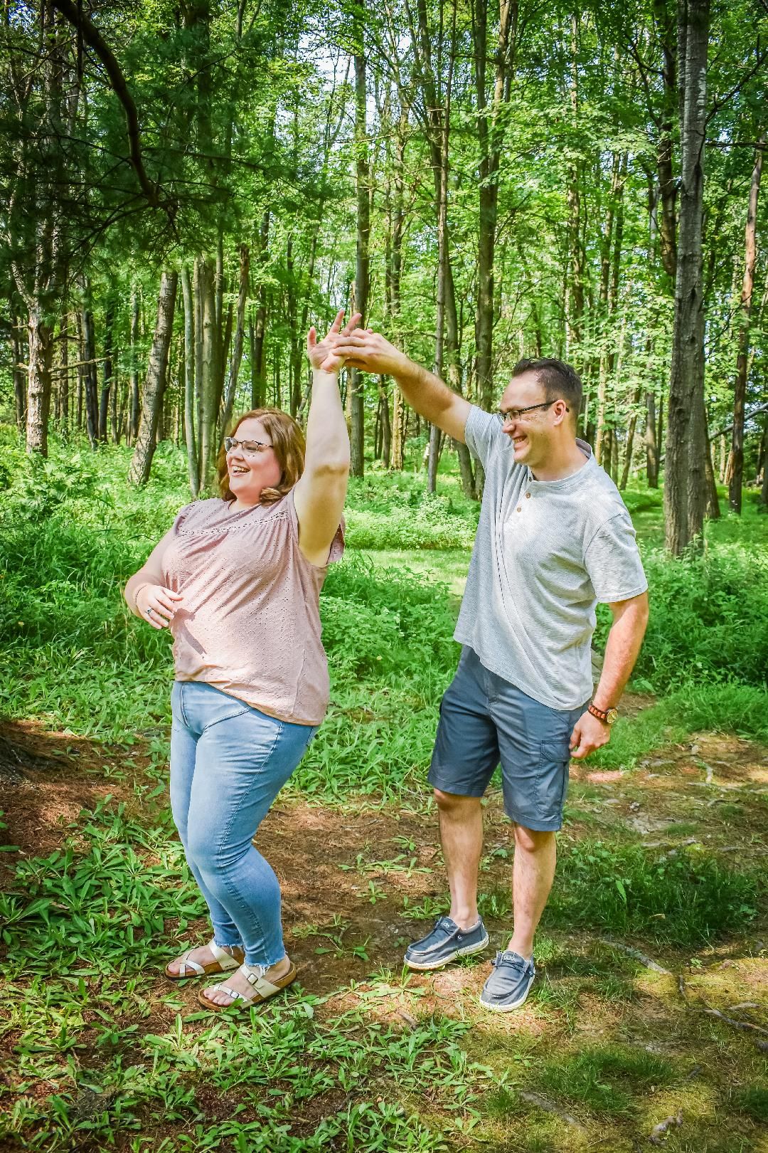 Husband and wife holding hands, laughing, and dancing in an outdoor setting filled with nature and green leaves. Blue Onion Photography.