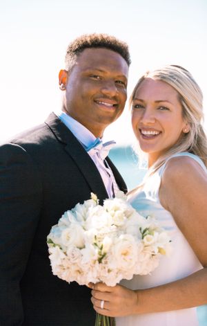 Wedding couple poses with white floral bouquet against bright sky background.