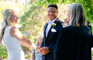 Wedding celebration outdoors with guests gathered under green summer foliage.