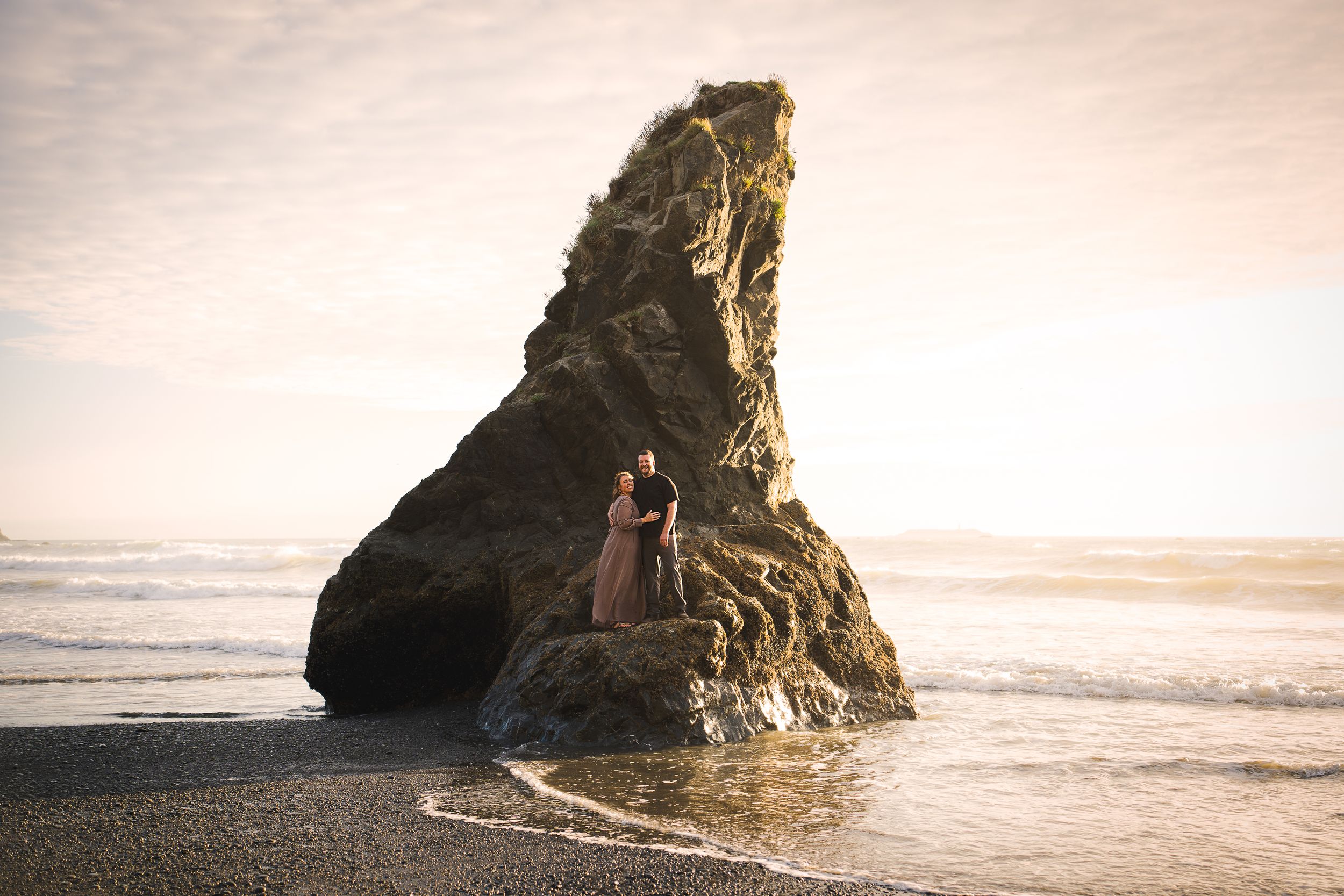An engagement photoshoot of a couple standing on a large rock formation at sunset on a beach along the Washington coast.