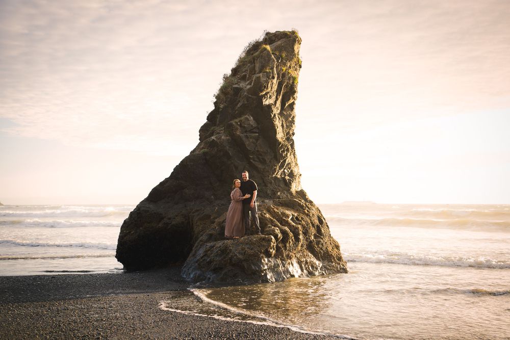 An engagement photoshoot of a couple standing on a large rock formation at sunset on a beach along the Washington coast.