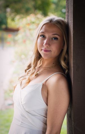 Portrait of a senior student, young woman, in a white dress leaning against a wooden post outdoors in natural lighting.