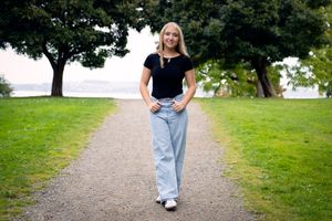 Senior student, young woman, in a black t-shirt and light blue jeans walks along a gravel path surrounded by green grass and trees.