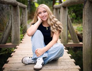 A senior student, young woman, in light jeans and black top sits casually on a wooden footbridge during an outdoor photo session.