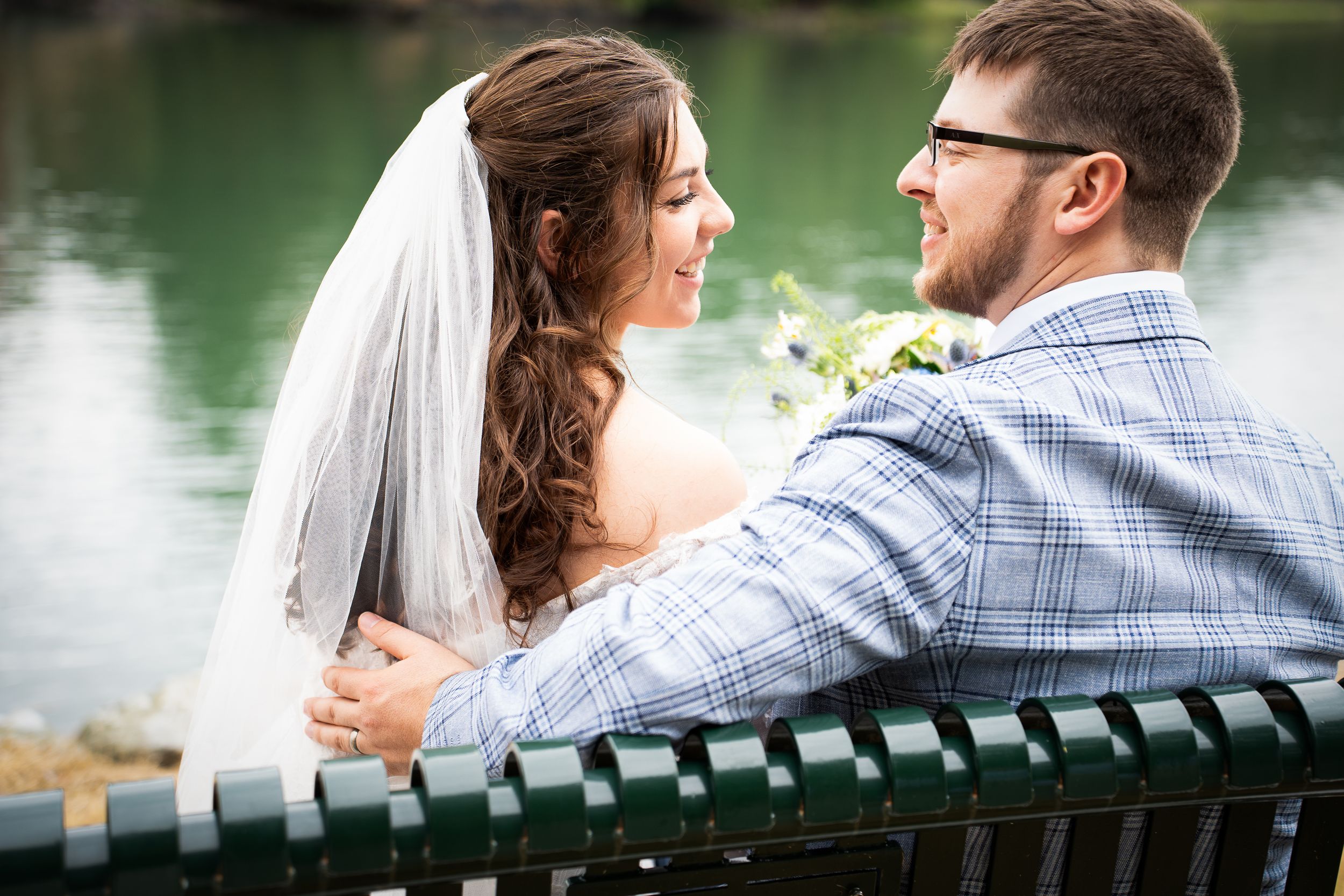 A romantic couple sits together on a wooden bench beside a serene lake on their wedding day.