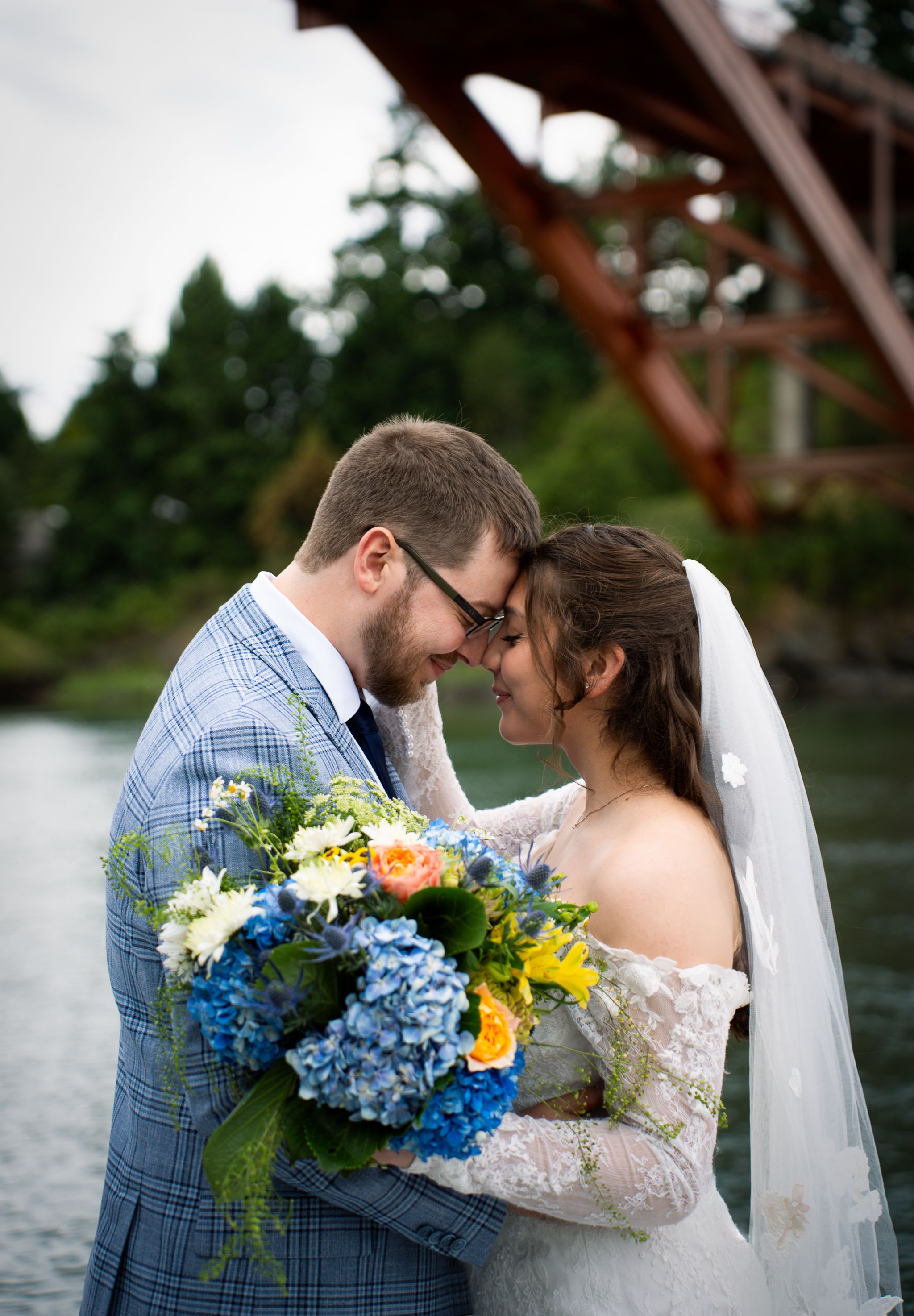 Newlywed couple shares an intimate moment with blue hydrangeas and yellow sunflowers in wedding bouquet by the water.