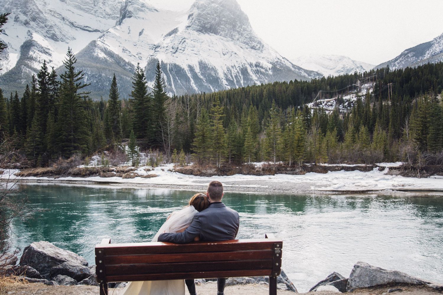 Person sitting on bench overlooking turquoise mountain lake with snow-capped peaks in background.