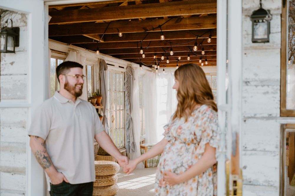 Expecting couple holds hands in rustic barn doorway, man with tattoos and pregnant woman in floral dress.