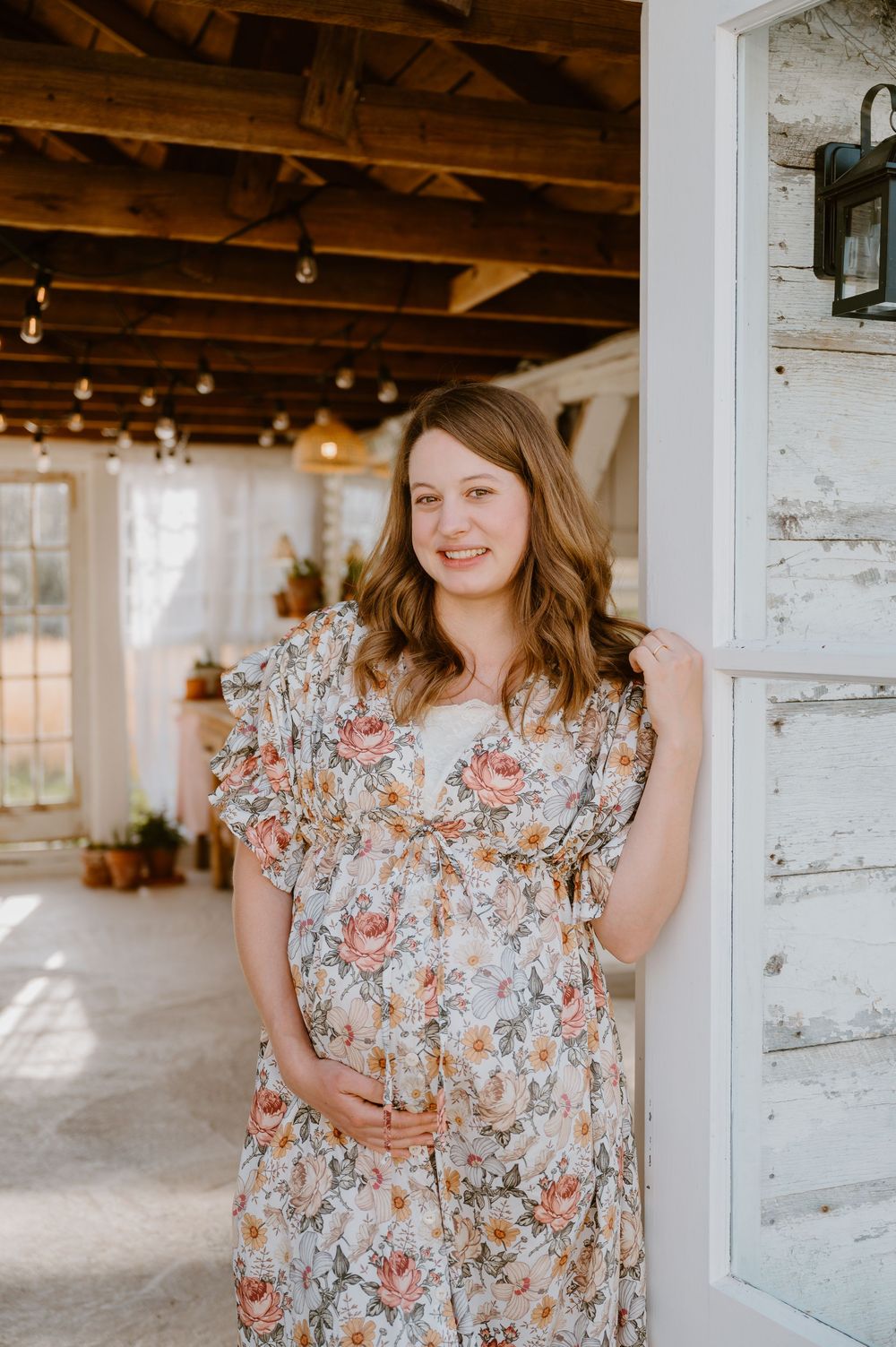 Pregnant woman in floral dress smiling by white barn door during maternity photo session.