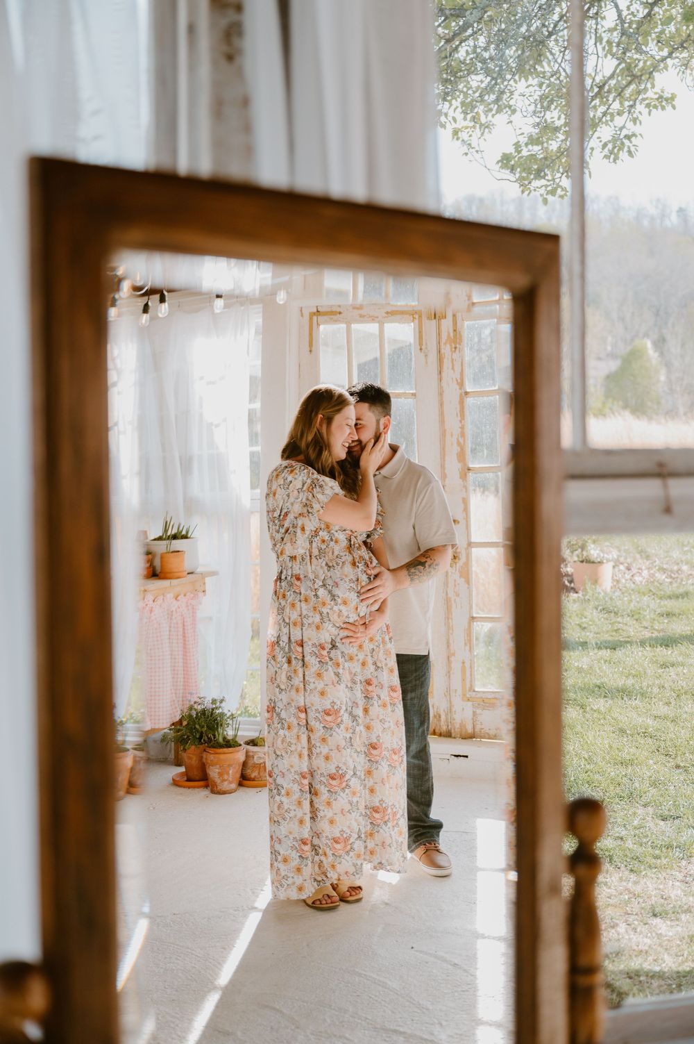 Pregnant couple shares a tender kiss in a sunlit boho studio, woman in floral maxi dress with hands cradling baby bump.