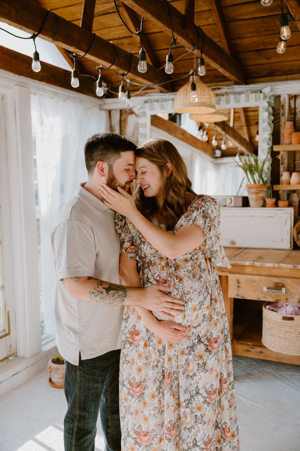 A couple shares a tender moment in a rustic boho indoor setting, with the woman in a floral dress cradling her baby bump.