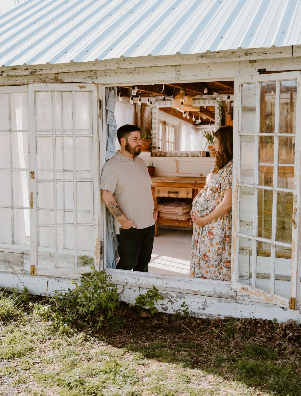 Pregnant couple standing in doorway of rustic white greenhouse building with French doors and tin roof.