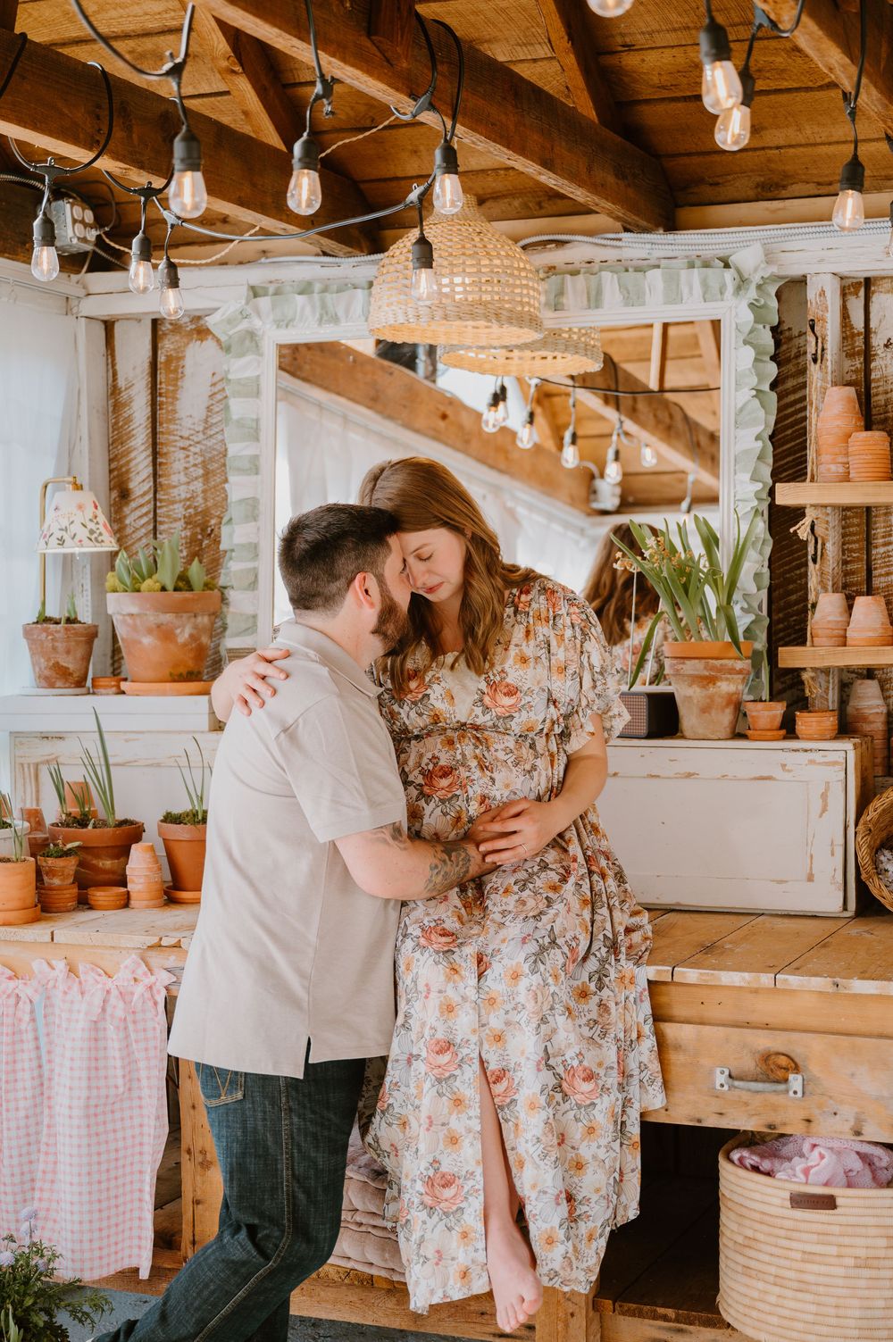 Couple sharing tender moment embracing baby bump in rustic greenhouse with string lights.