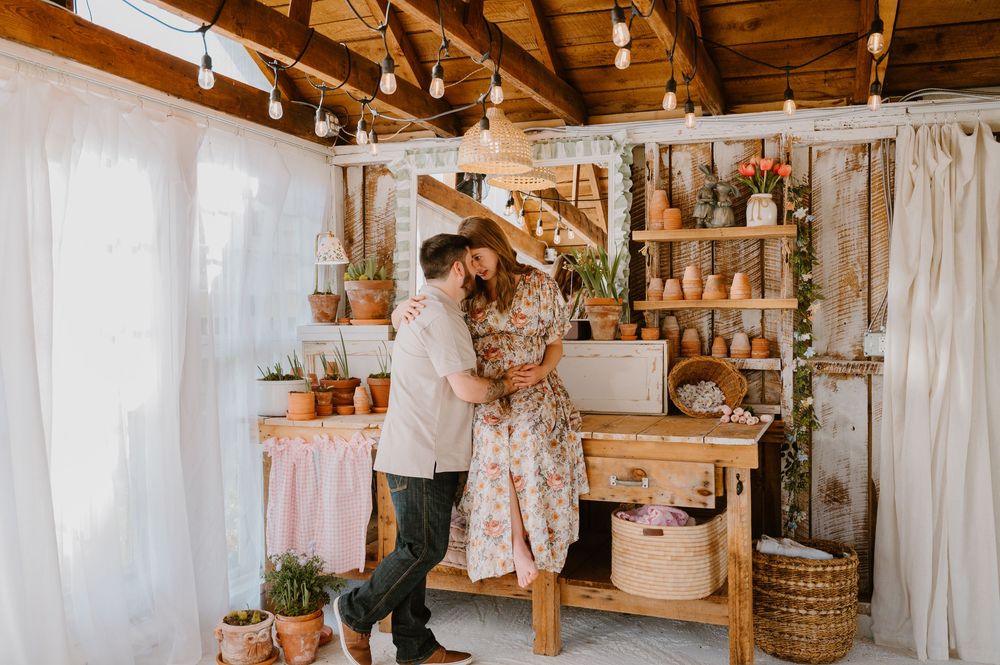 Couple embracing romantically in a rustic boho workshop with wooden shelves, potted plants, and warm string lights.