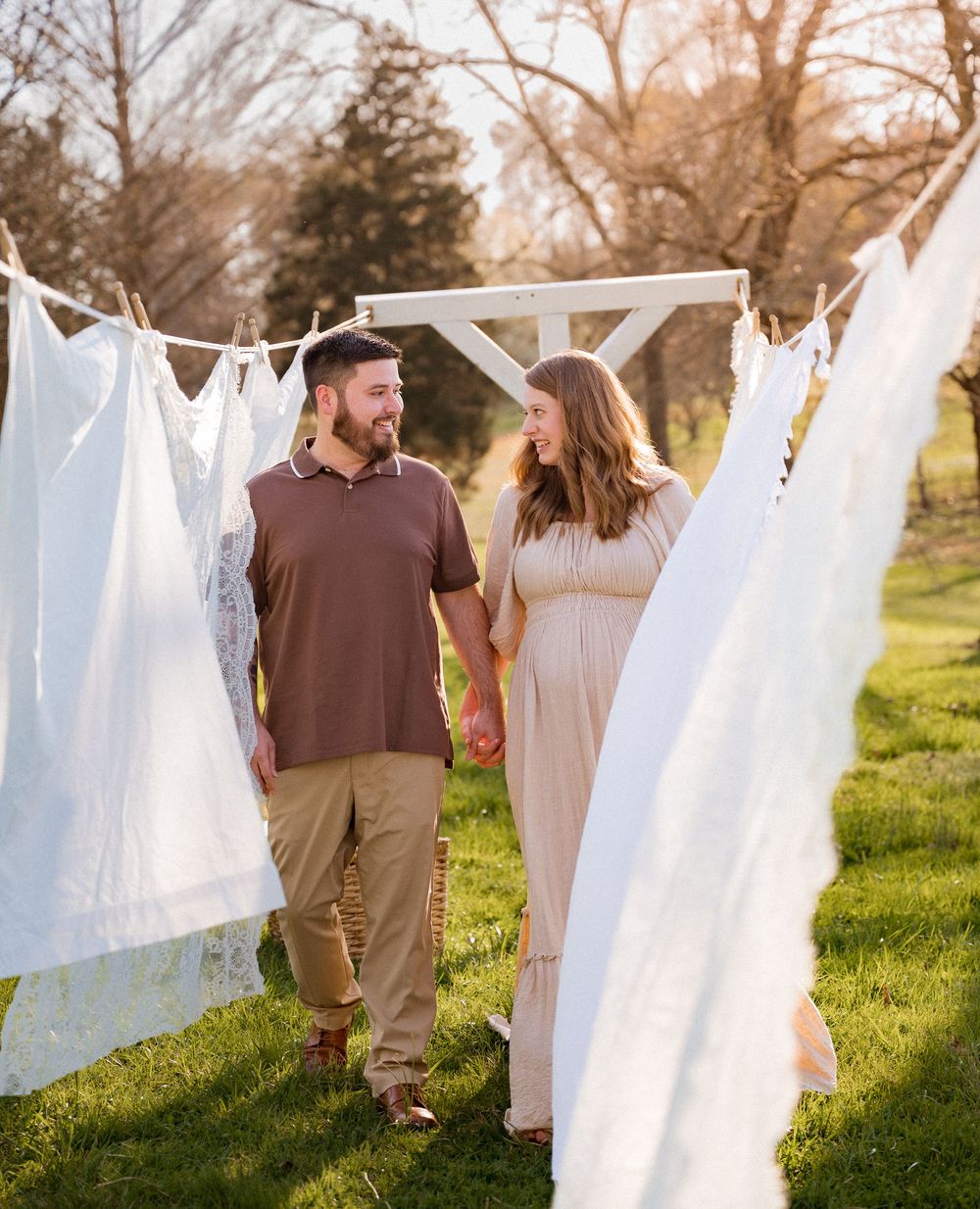 Couple holding hands walking between white linens on a clothesline outdoors during golden hour.