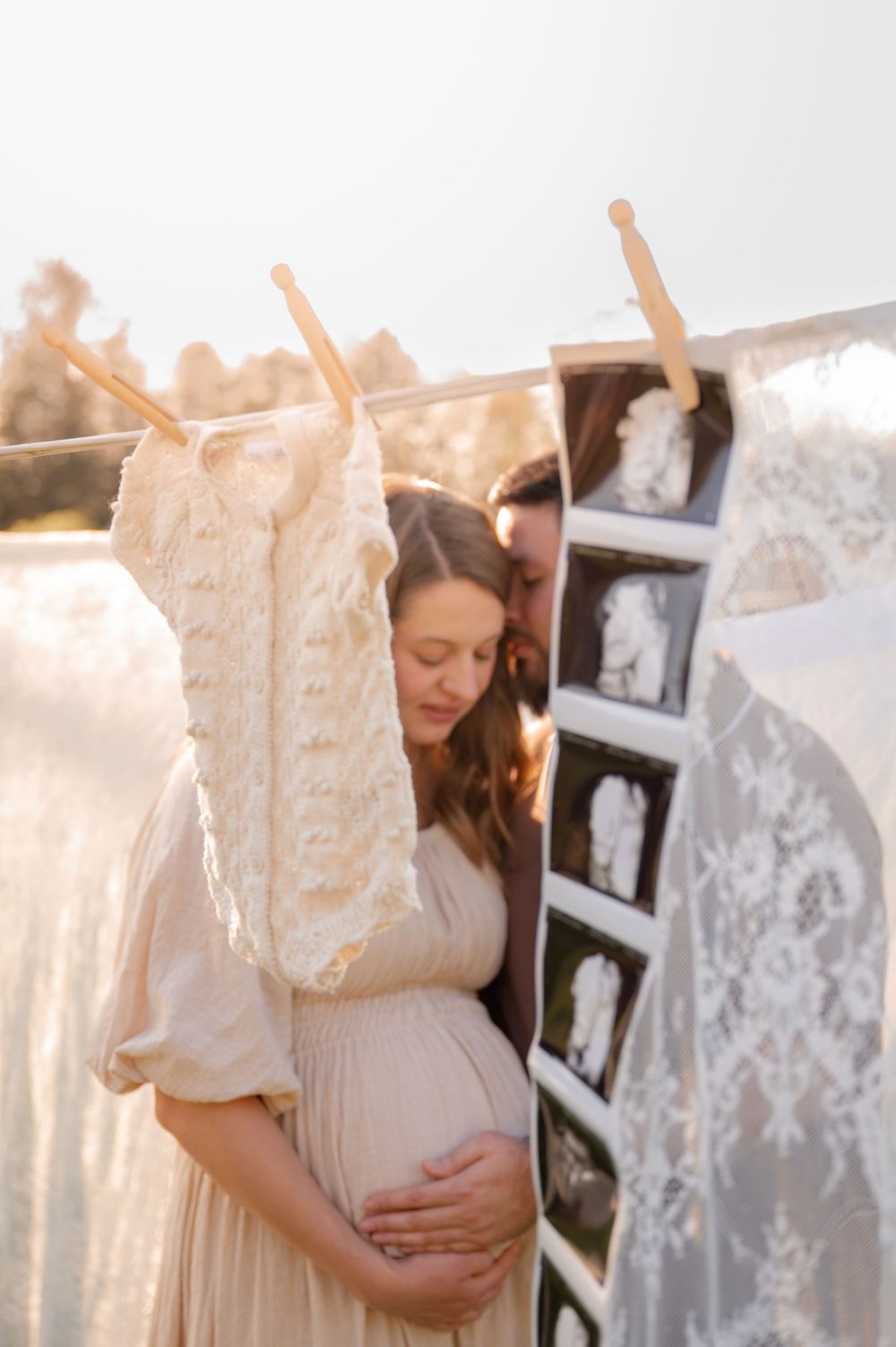 Pregnant woman in beige dress holds belly beside clothesline with baby clothes and ultrasound photos during golden hour.