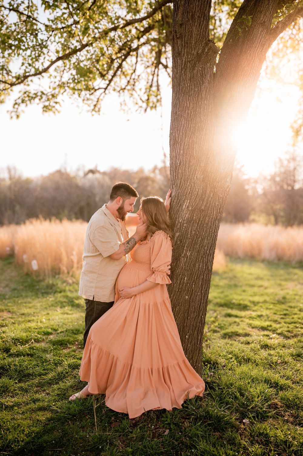 A pregnant couple embraces near a tree at golden hour, the woman wearing a flowing peach dress in a sunlit field.