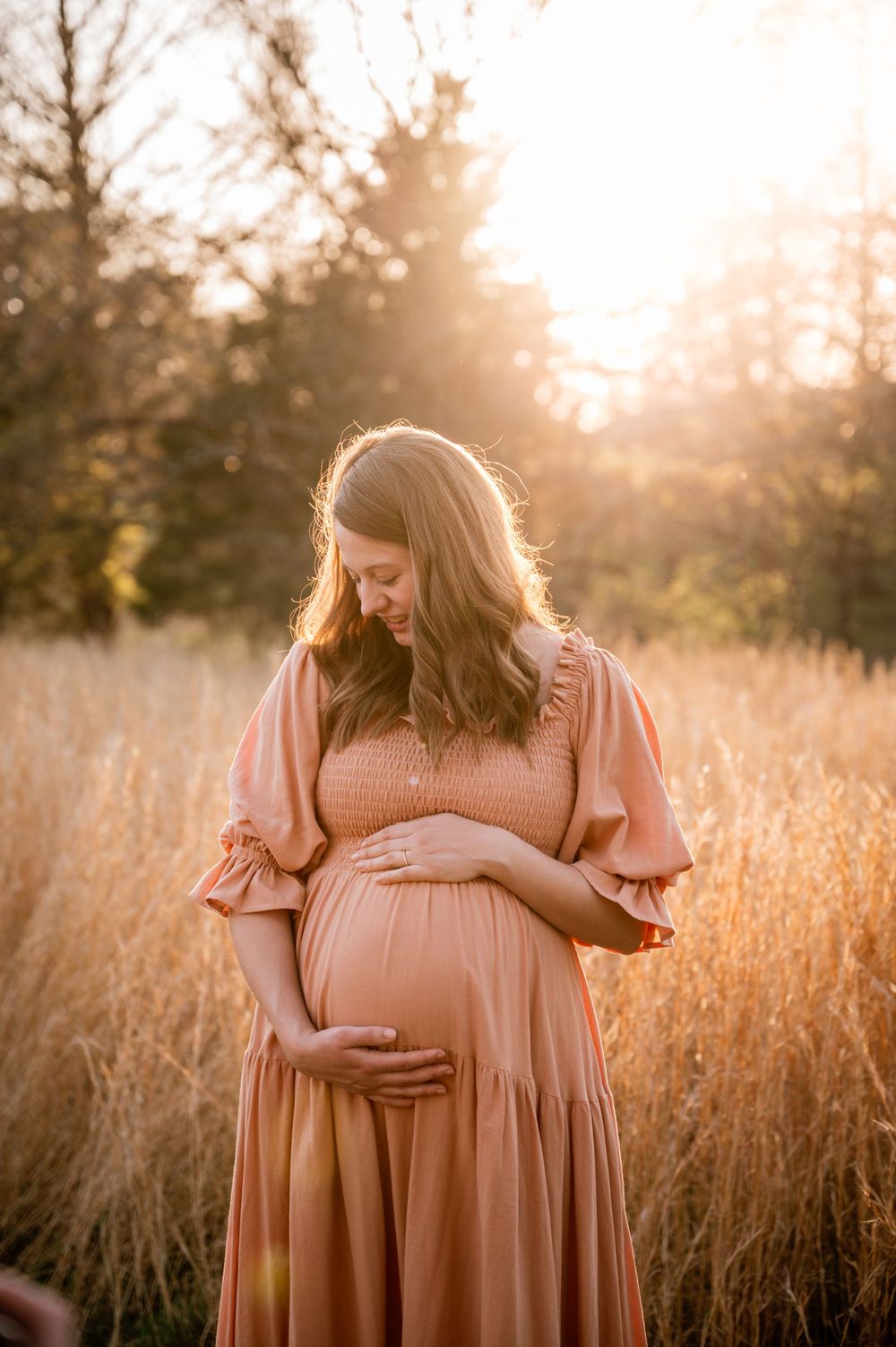 Pregnant woman in a peach dress cradling her baby bump in a golden field at sunset.