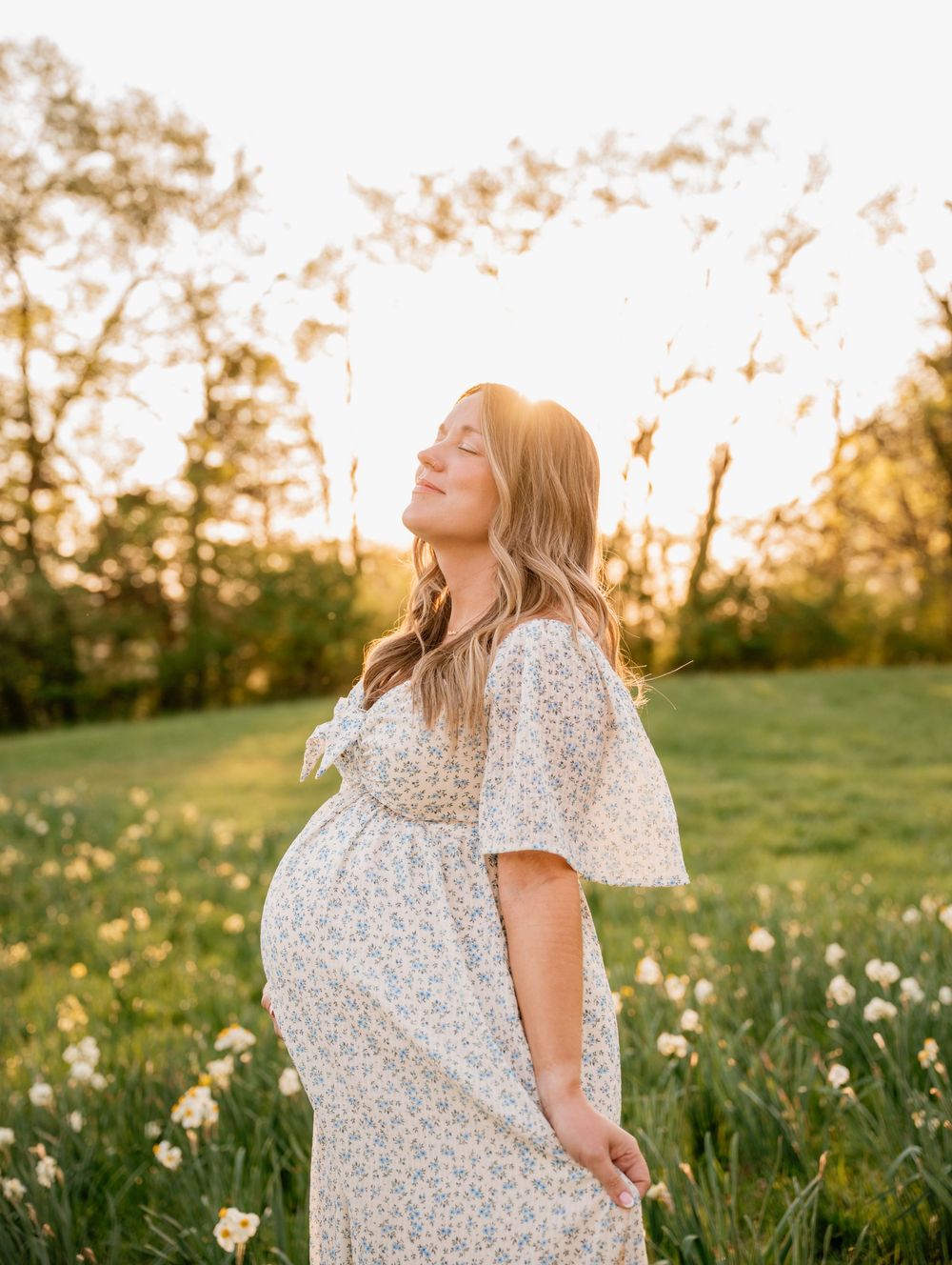 Pregnant woman in floral dress standing in sunny meadow with wildflowers, golden hour light, serene maternity photo.
