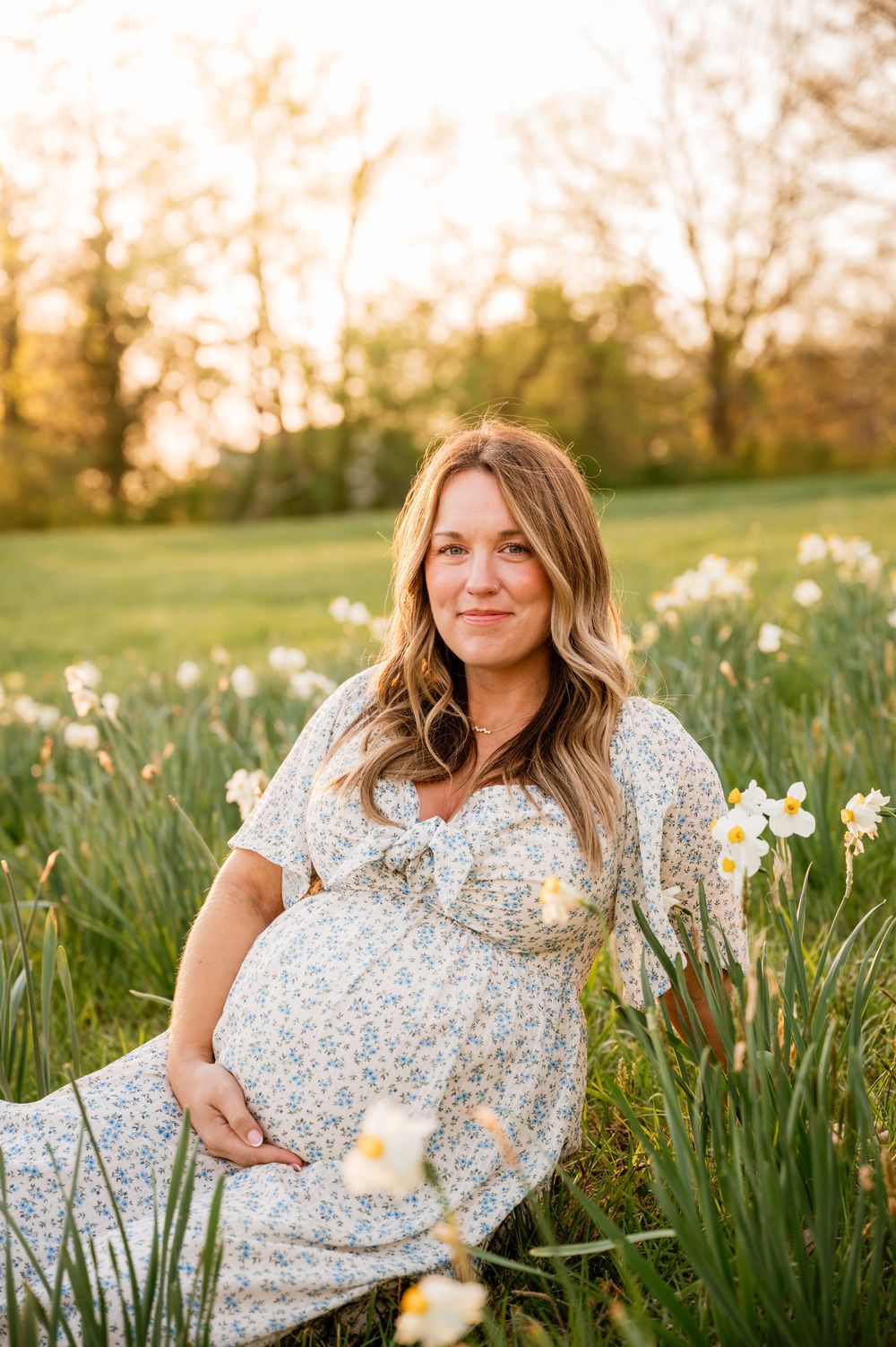 Pregnant woman in floral dress sitting among white daffodils in a sunlit field during golden hour maternity photoshoot.