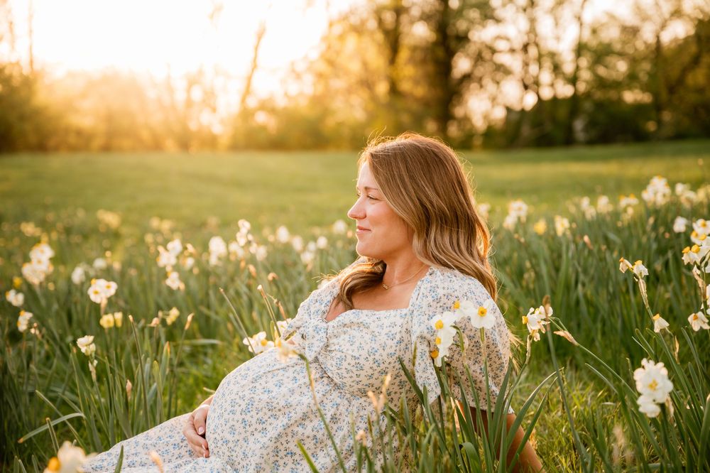 Pregnant woman in floral dress sitting in a sunlit field of white daffodils during golden hour maternity photoshoot.