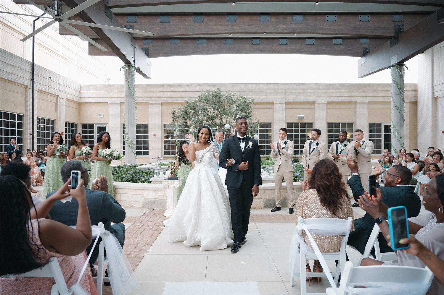 Wedding ceremony guests capture a joyful wedding procession down an aisle with their phones at an elegant outdoor venue.