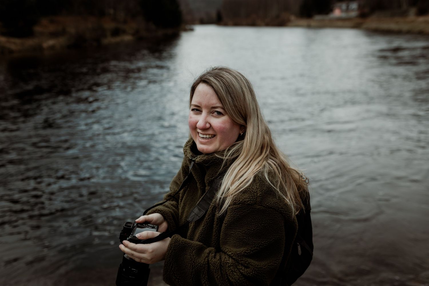 A winter portrait by a scenic river in Scotland holding a camera.