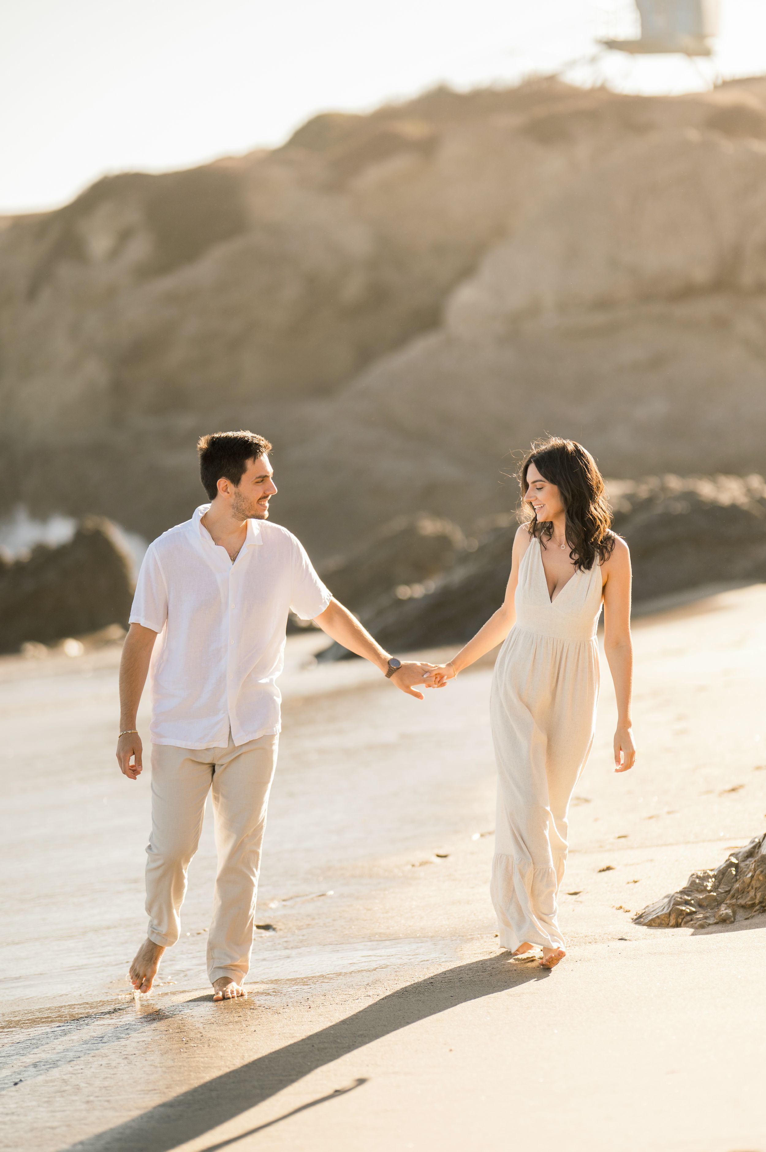 A couple walking hand in hand on a sandy beach during golden hour.