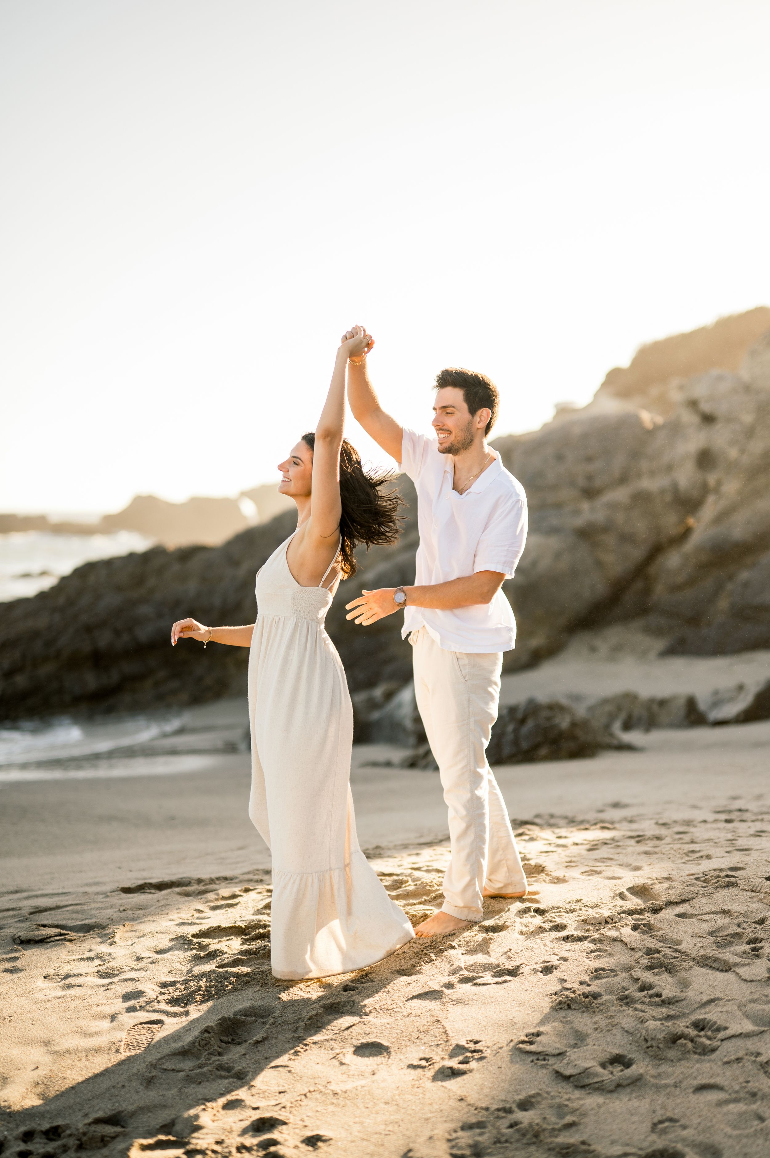 A romantic couple in white attire dancing together on a beach at sunset with rocky cliffs in the background.