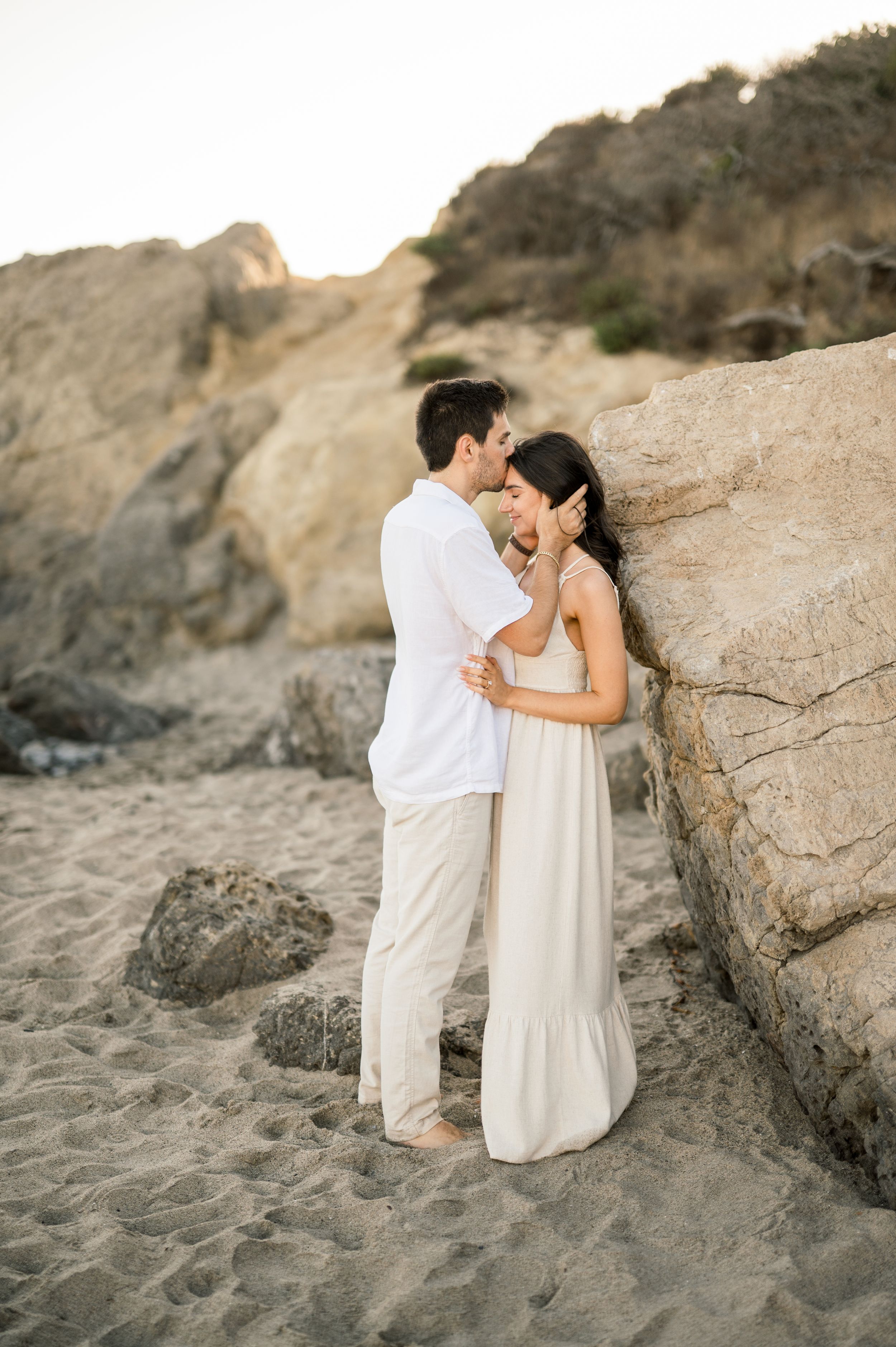A couple in white clothing sharing a tender moment against coastal rocks during golden hour.