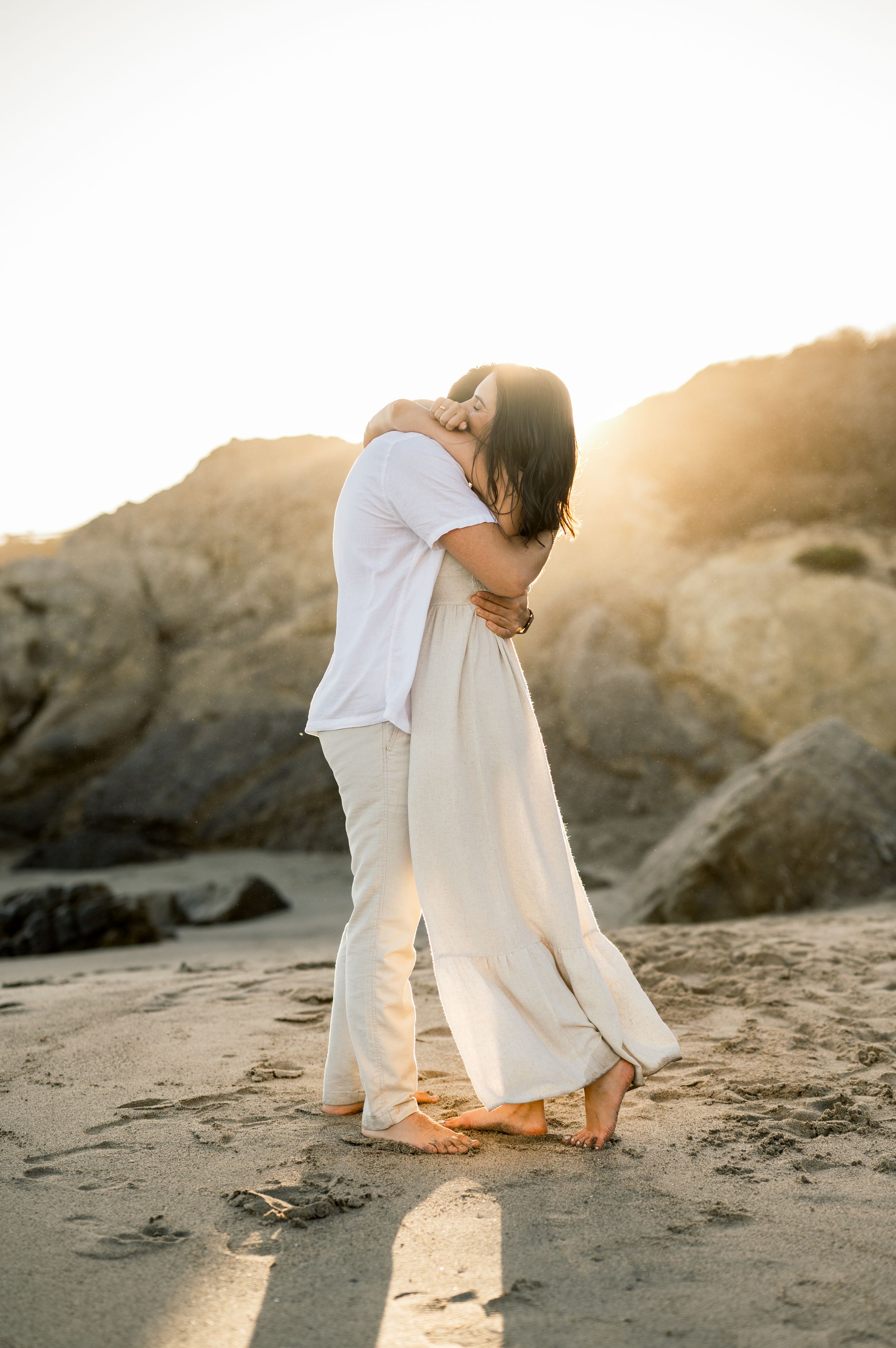 A couple in coordinating white outfits embracing on a sandy beach during sunset.
