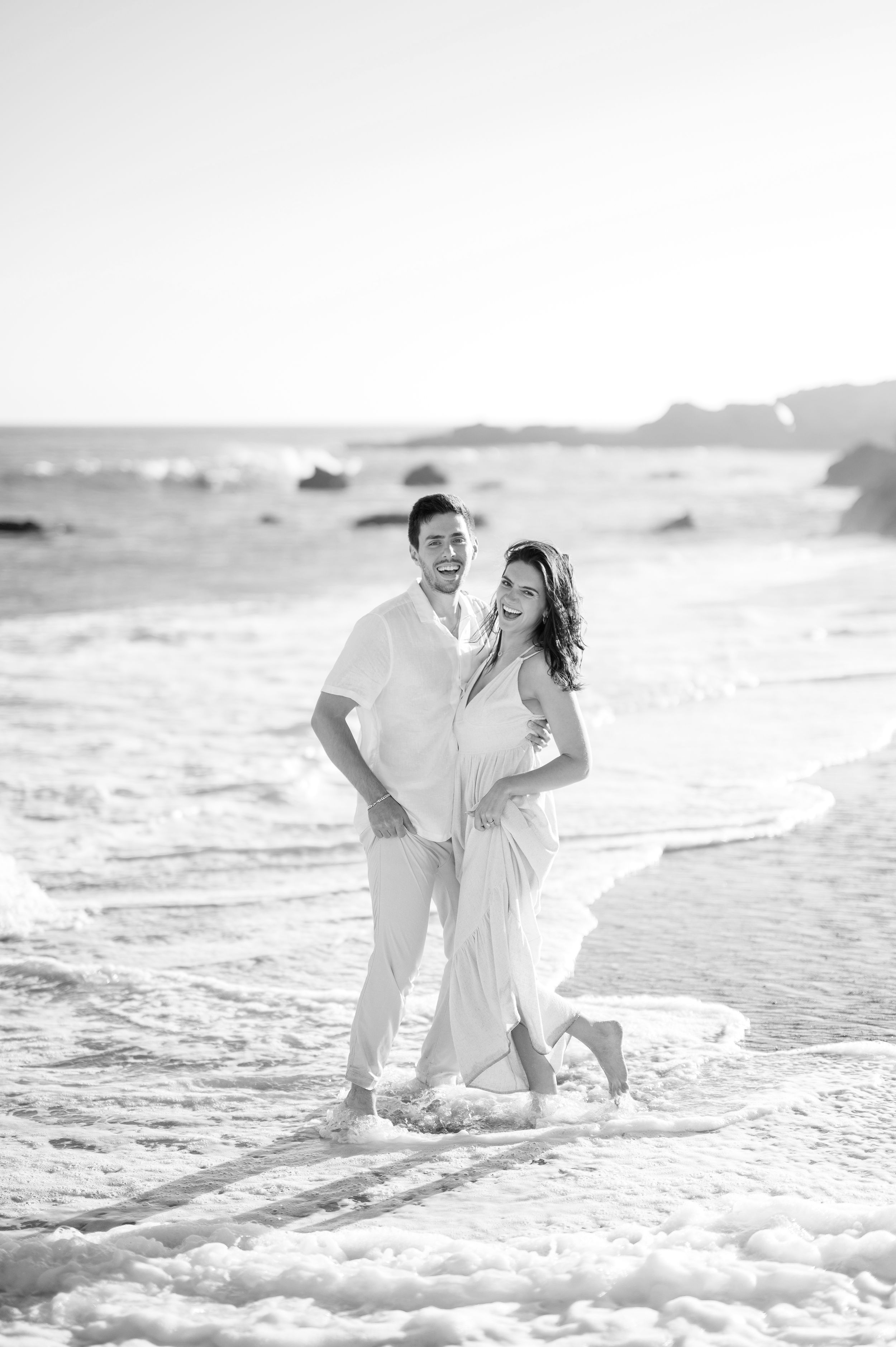A joyful couple in white clothing playing in the ocean waves during a beach photoshoot.