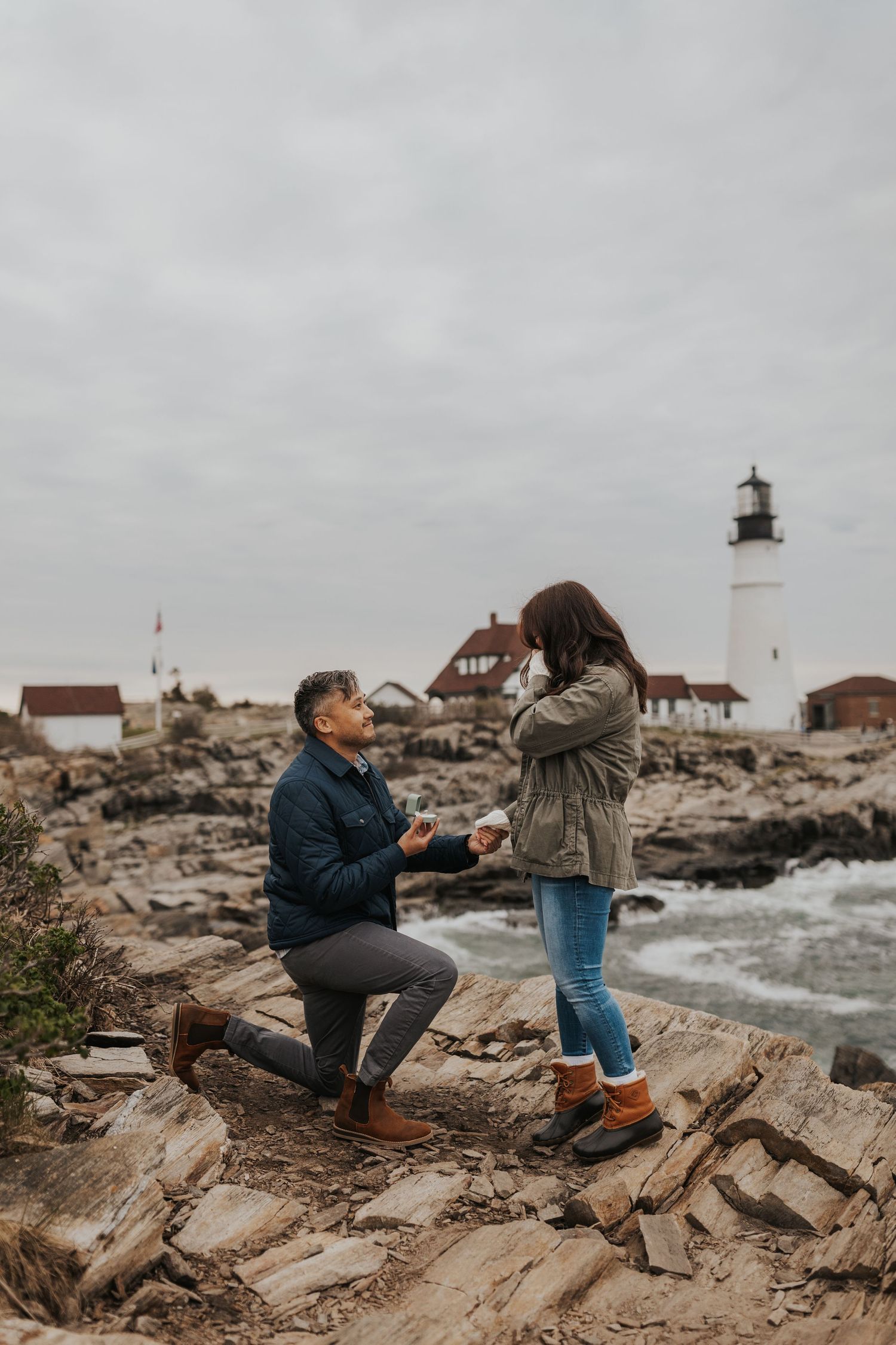 A surprise proposal scene in front of a lighthouse with rocky coastal backdrop.