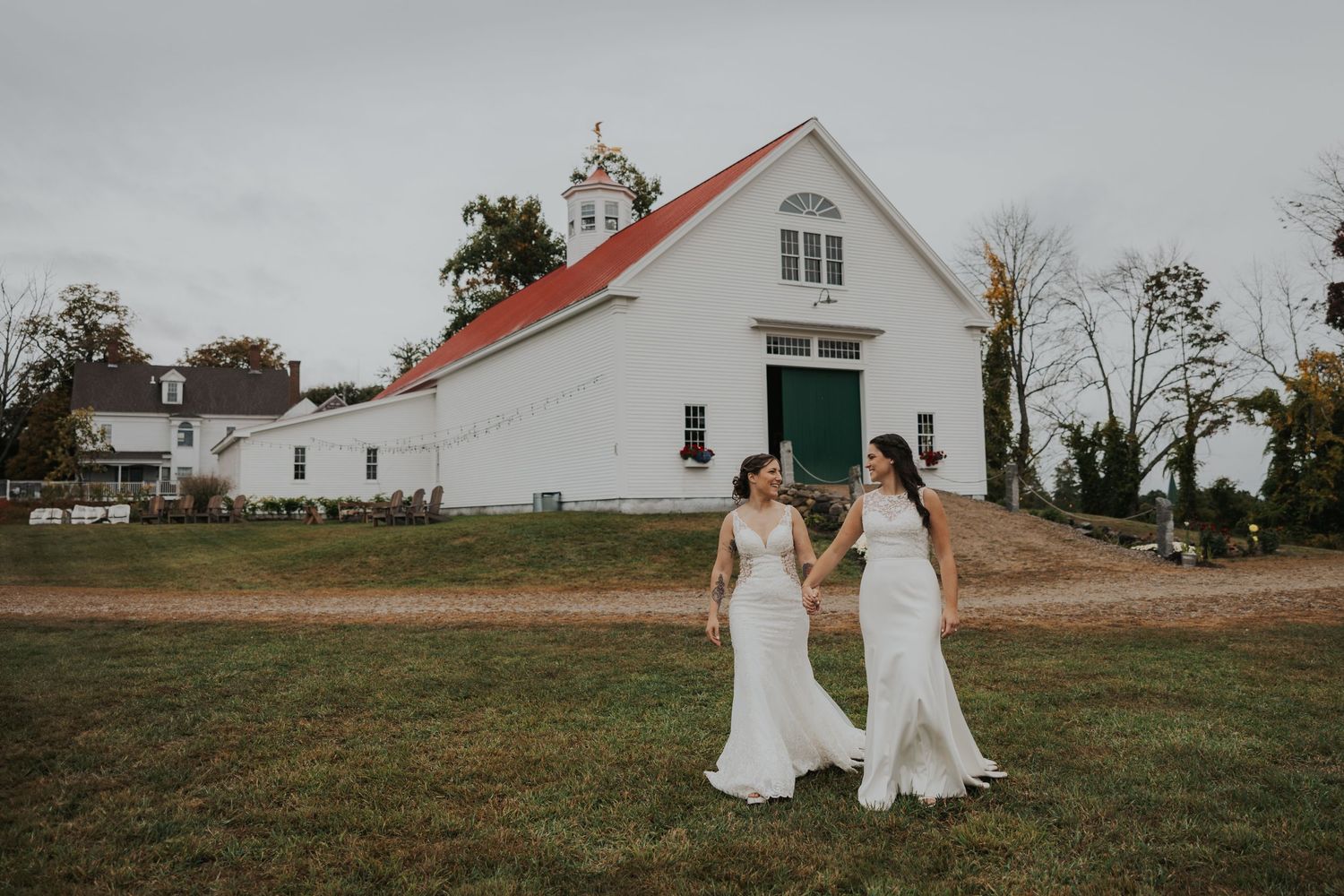 Two brides walking together in white dresses with rustic barn in background in New Hampshire.