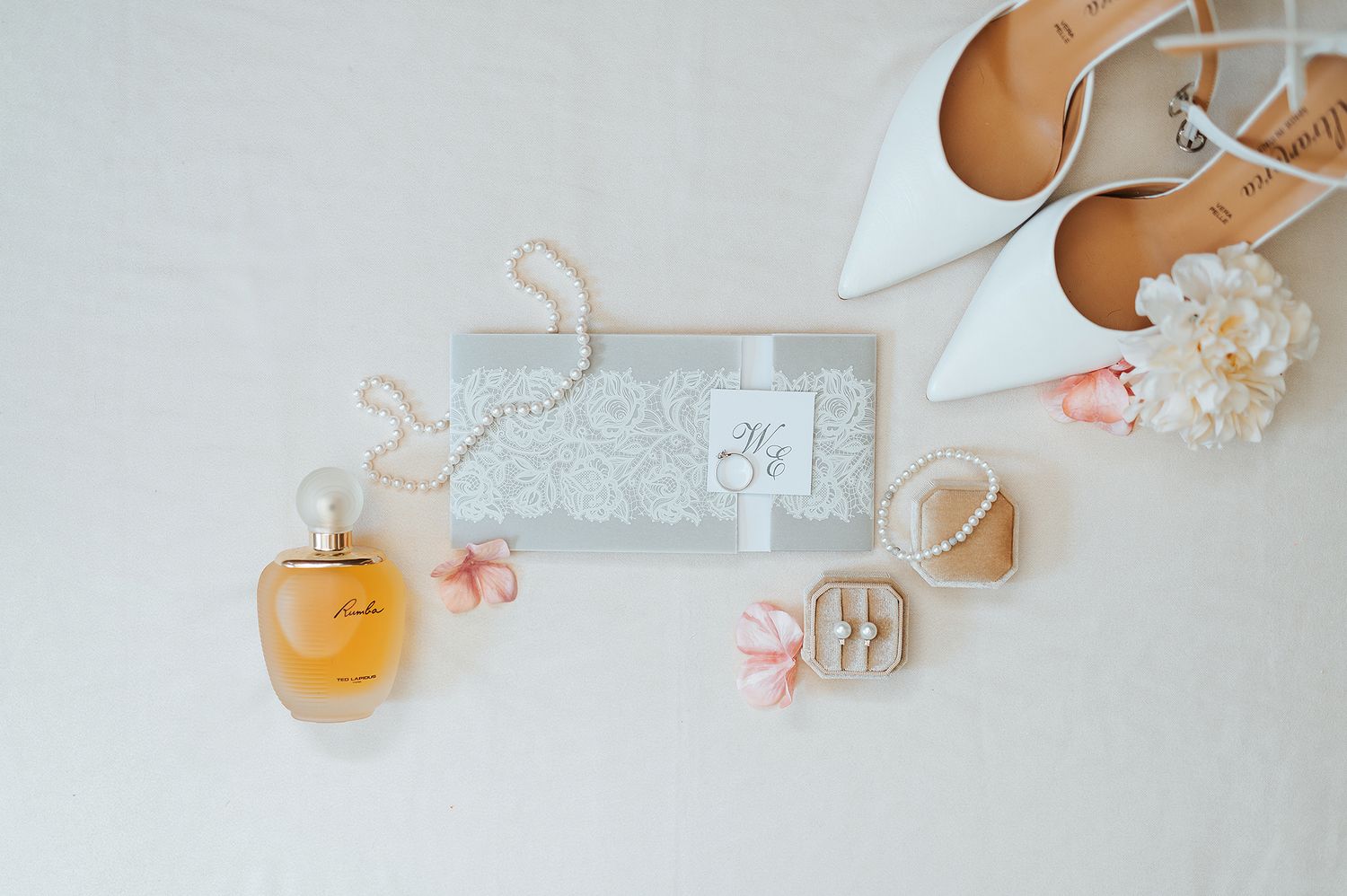 Elegant bridal flat lay with white heels, pearl necklace, perfume, rings, and floral invitation.