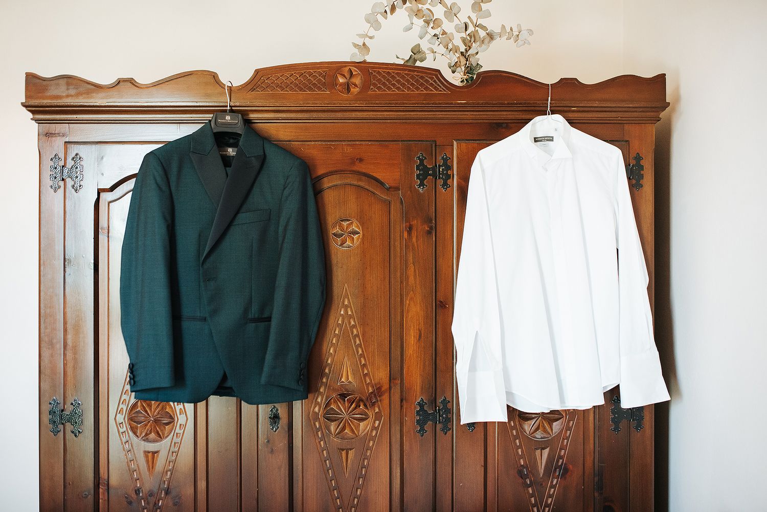 Dark green groom suit jacket and white dress shirt hanging on ornate wooden wardrobe.