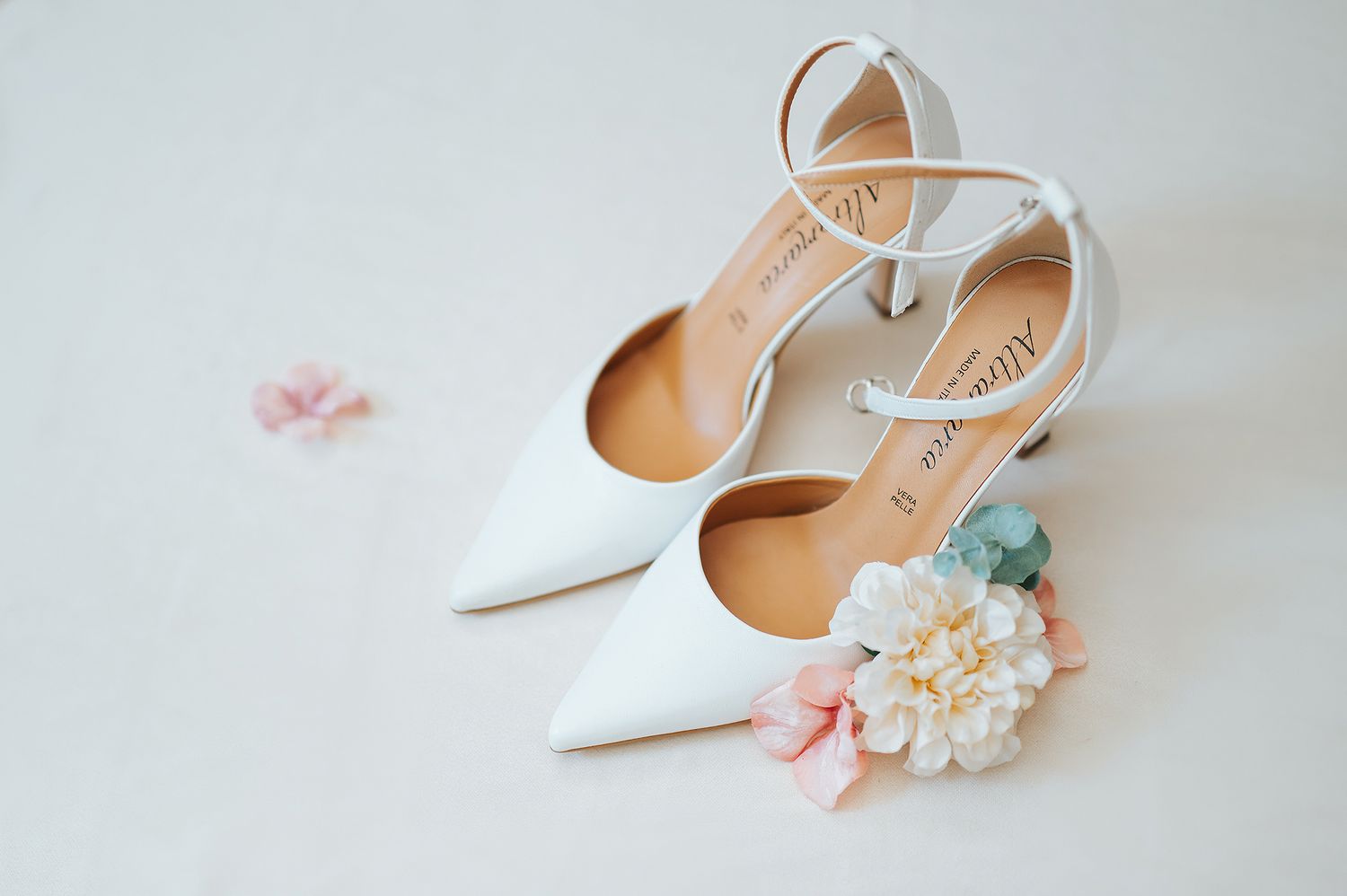 White pointed-toe bridal heels with ankle straps displayed beside delicate flowers on white surface.