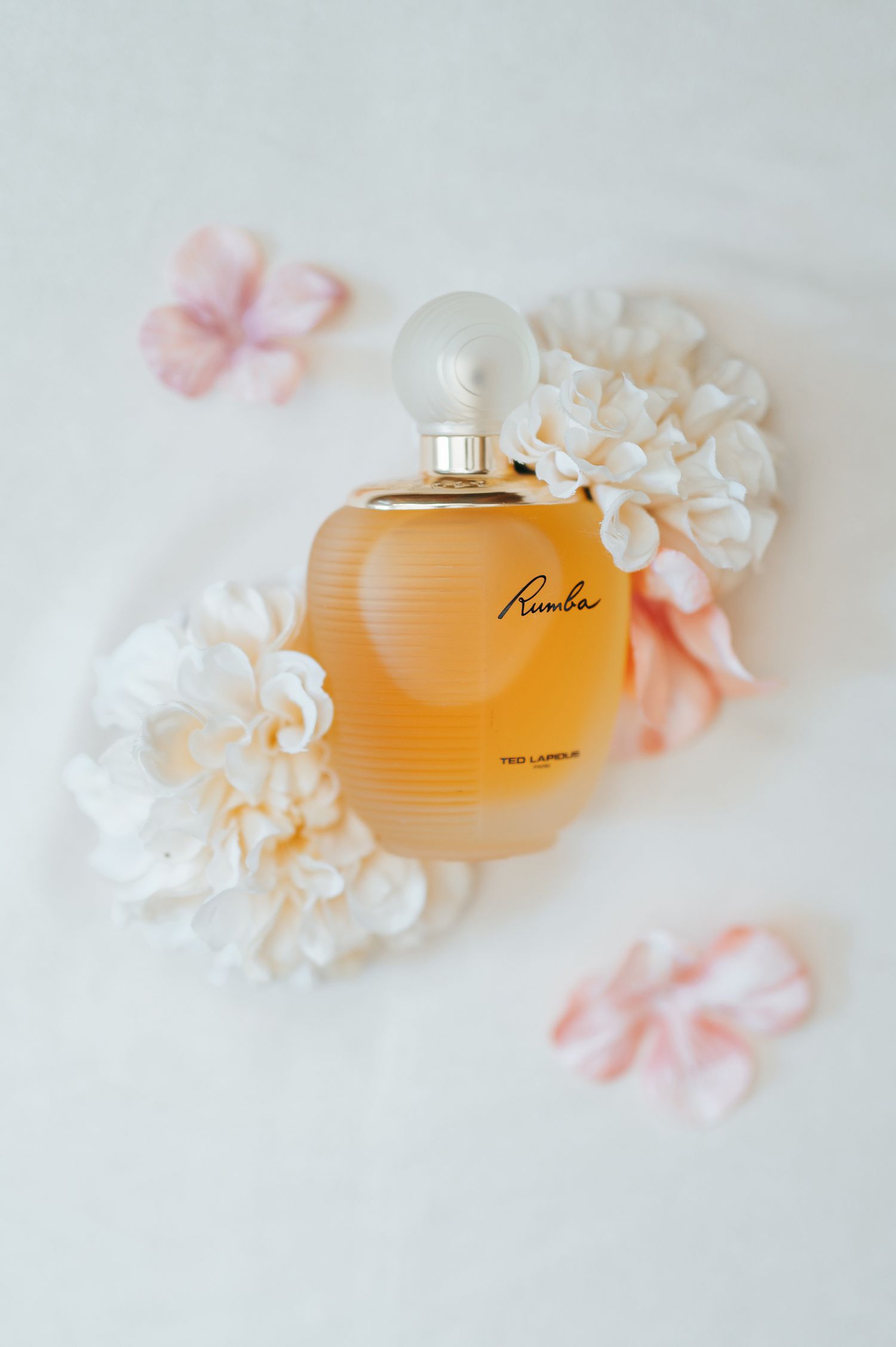 Rumba by Ted Lapidus amber perfume bottle surrounded by white and pink flowers on white background.