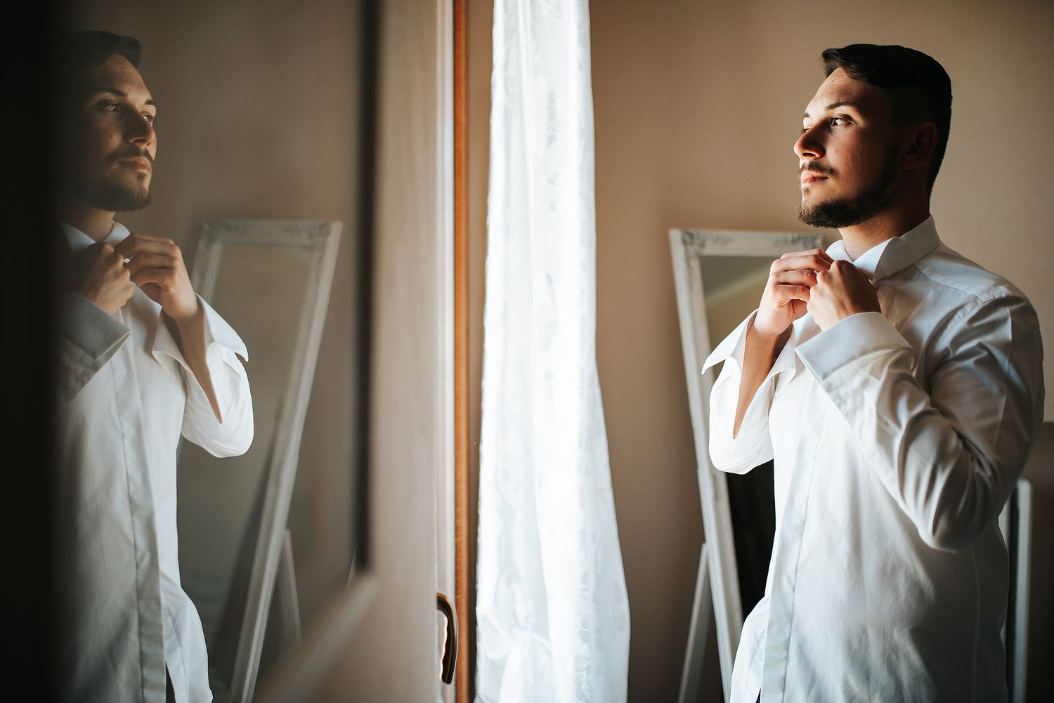 Groom in white shirt adjusting collar while looking in mirror before wedding ceremony.