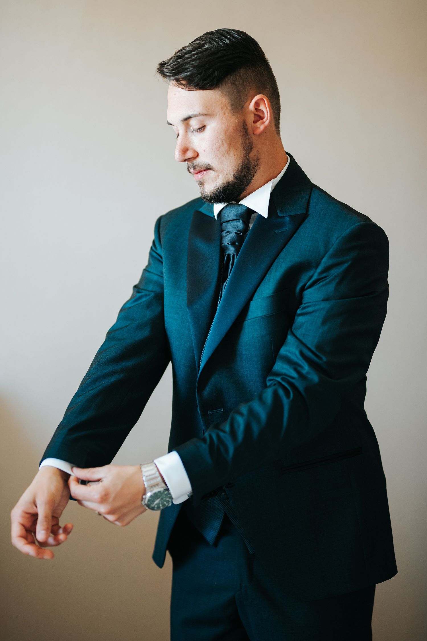 Stylish groom in dark teal suit adjusting cufflinks while wearing a watch before wedding.
