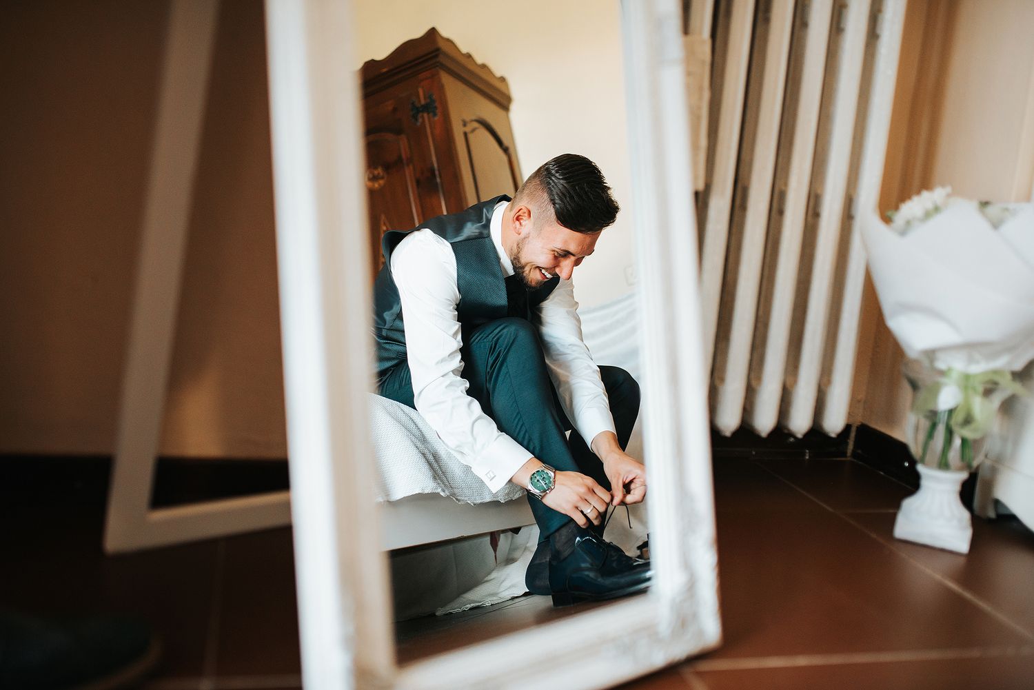 Mirror reflection of groom in vest and white shirt tying his shoes before wedding.