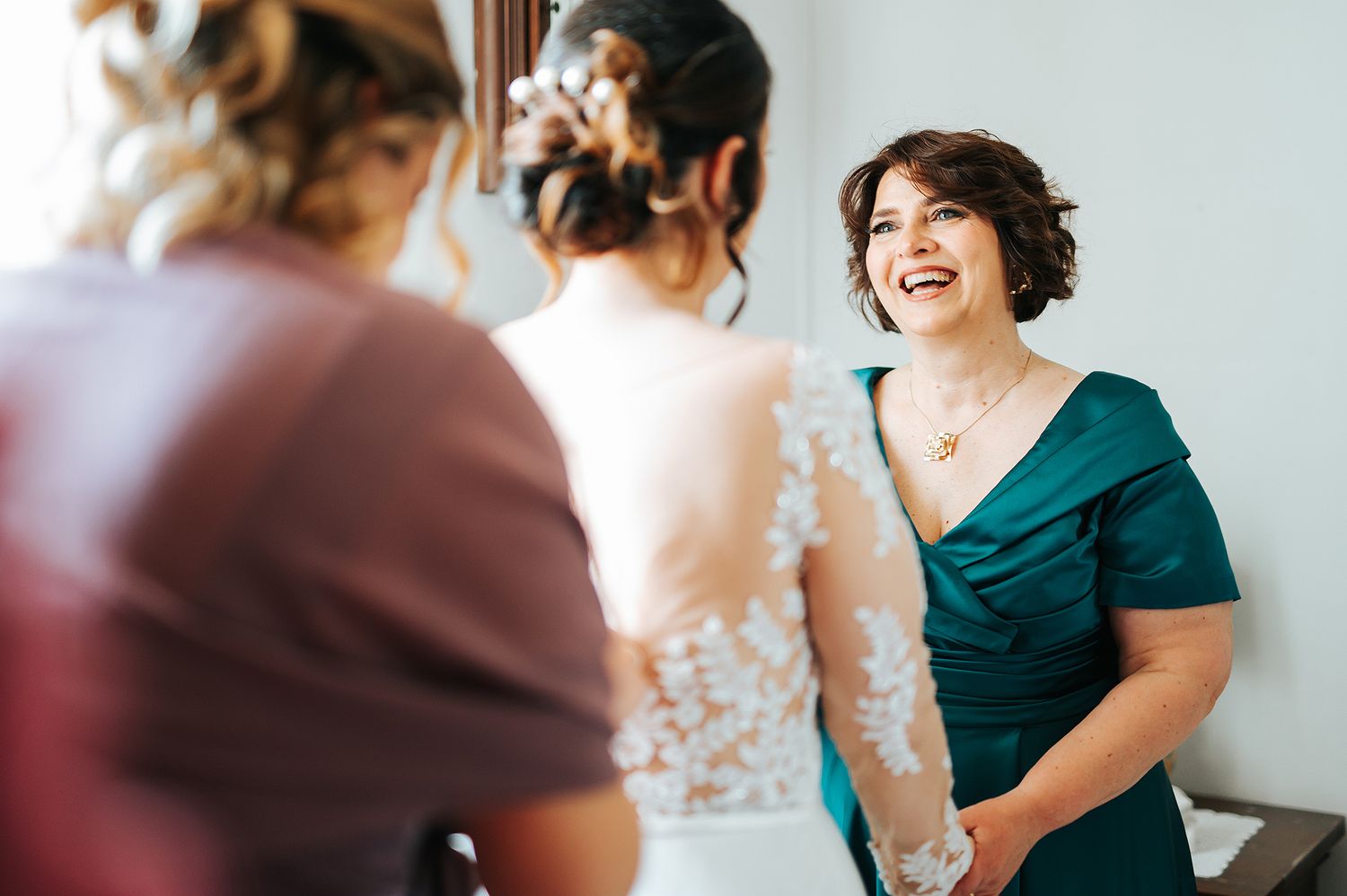 Joyful woman in teal dress laughing with bride during wedding preparation.
