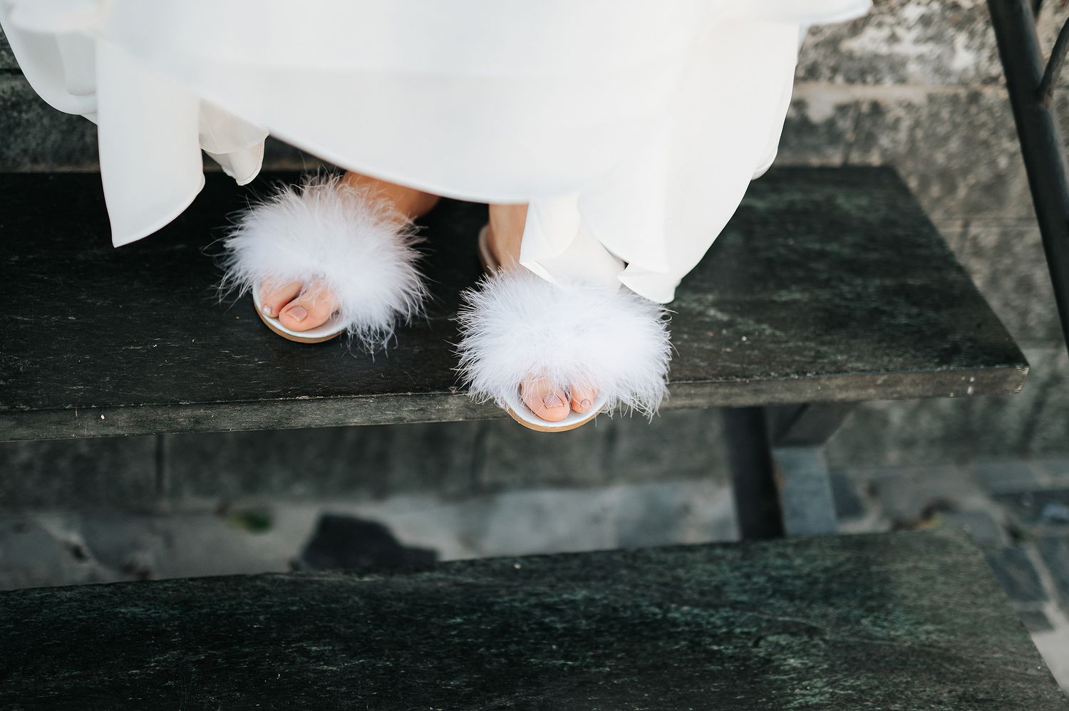 Bride wearing fluffy white feather wedding shoes peeking under bridal gown on steps.